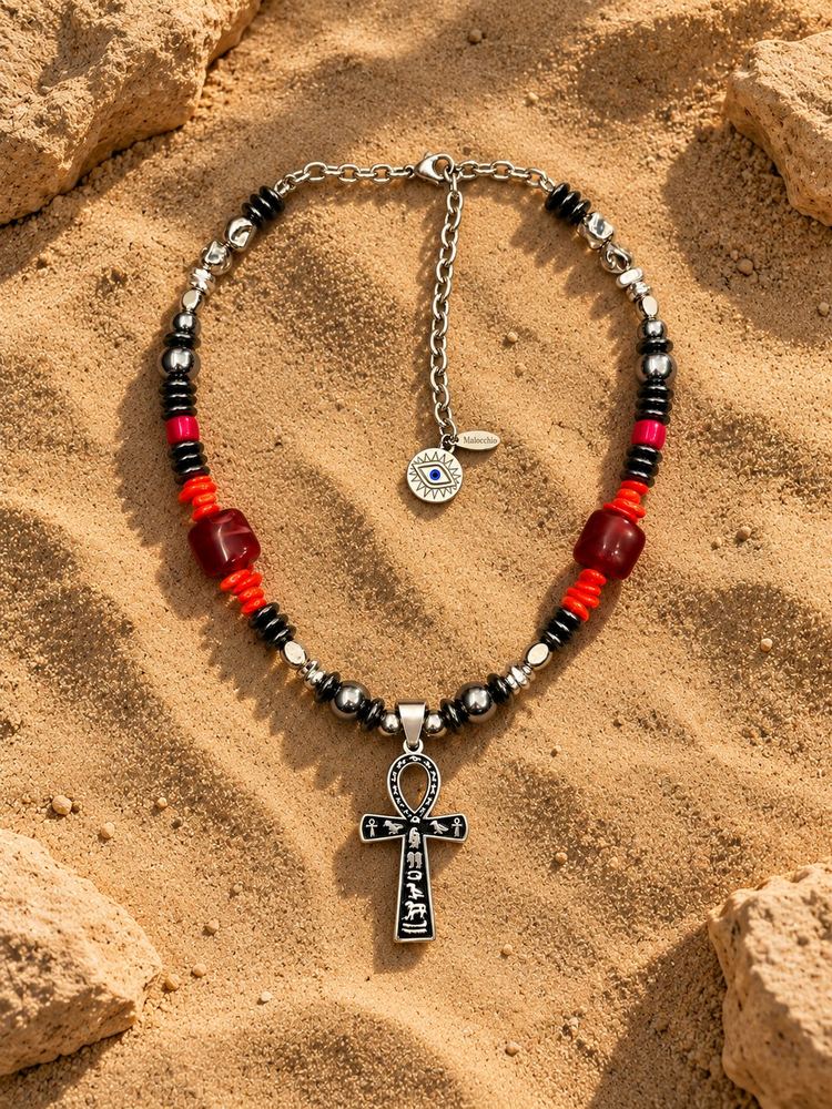 Collana MALOCCHIO «ANKH — SCARLET»: perle scure, dettagli metallici, accenti rosso fuoco e ciondolo ankh, sulla sabbia