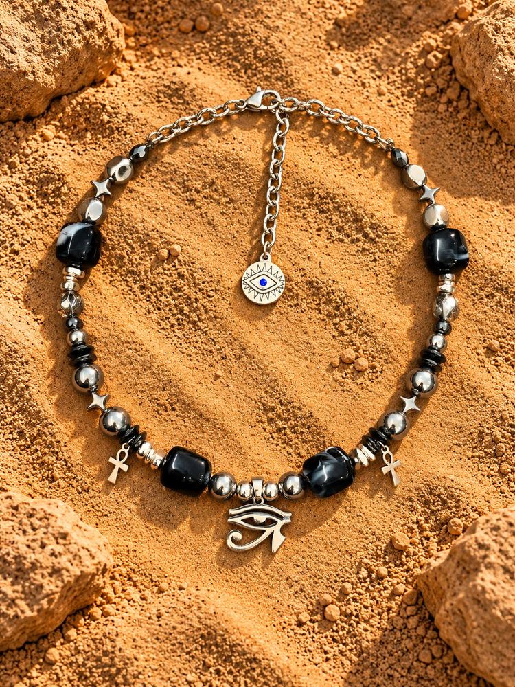 Collana MALOCCHIO «HORUS — OBSIDIAN»: perle nere marmorizzate, due piccoli ankh e Occhio di Horus argentato, sulla sabbia